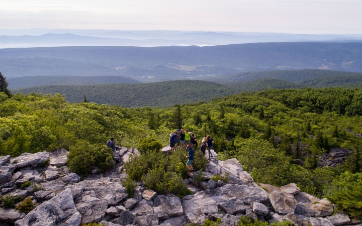 A group of backpackers take in the view of vast trees and mountains from atop a rocky outlook. 